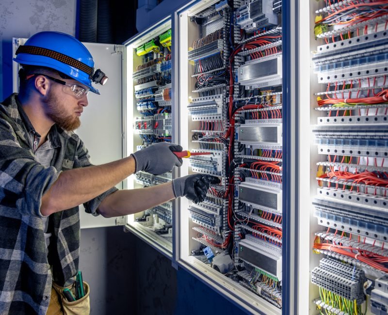 Male electrician working in switchboard with electrical connecting cable. Young adult electrical engineer in special clothes with flashlight on helmet in dark room with emergency lights in background. SSUCv3H4sIAAAAAAAAA41Py46DMAz8F59BSgBtWX6l2kNIDEQNBCVOqwrx7+sA1V43J3vGmccG1kAHsmmEkTdRai1FKSUOZVvJtmxqMVRGDxoVQgFGEUIn67oVX7WoblX7XQt+BfQqWg3dBta5FCkosn6BjpmAi8FwjGgs+WCV420vIJKiFDHyN940a4/MHvtH775lnAOyeUx9Hvbin9hPAWrERb+z/p6DOFSH3Z2px4swzJe3pylHZBGfFgrvU8h5v6receNBuYgFTCpGPjAXwB/XYLVdxqsBF/fzOS+estORh7x+WHNZnaUny2j22SCtziuD5qJTP1uizxrwafH1d73v+y+94WEbsgEAAA==